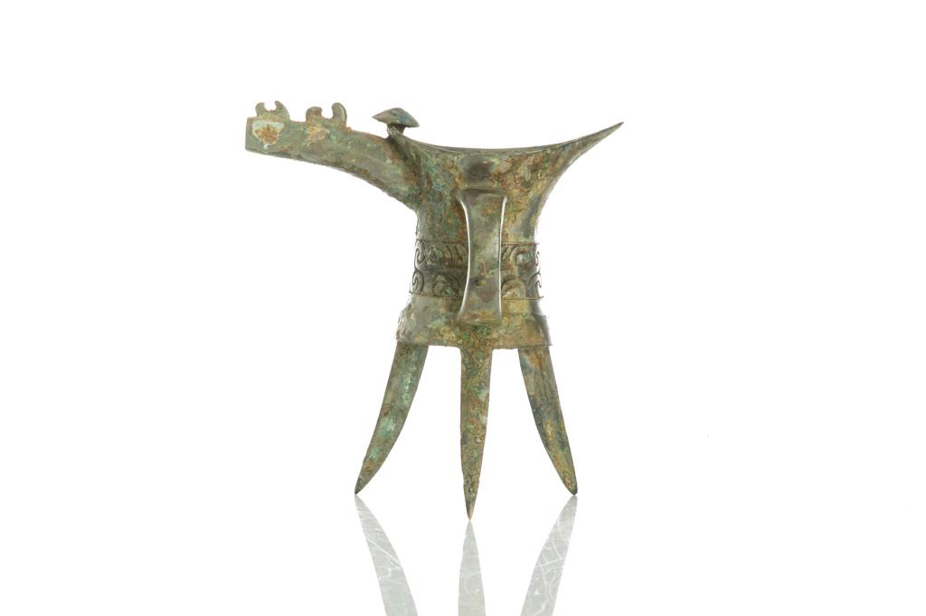 EARLY CHINESE BRONZE JUE CUP: Tripod legs supporting a cup with strap handle to one side and unusual spout. Taotie mask band to body. Possibly Erligang period to early Shang dynasty. H. 16.5cm (6 1/2 in.)