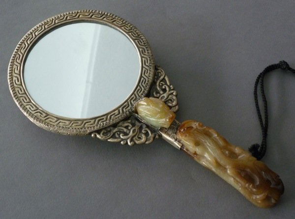 CHINESE HAND MIRROR WITH JADE BELT HOOK: Chinese metal hand mirror mounted with a green and brown jade belt hook as a handle. Length of mirror 23.5 cm (9 1/4 in), length of hook 12 cm (4 3/4 in).