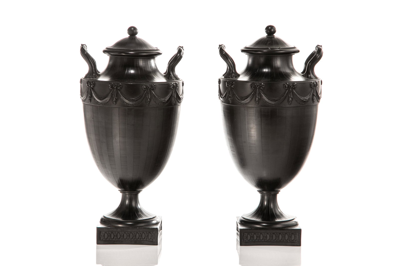 PAIR OF EARLY WEDGWOOD BASALT URNS: Two handled early English Wedgwood basalt urns. Each with Neoclassical swag decoration with low relief texture, topped with lid. Raised upon a square base with diaper pattern along the edge. Impressed