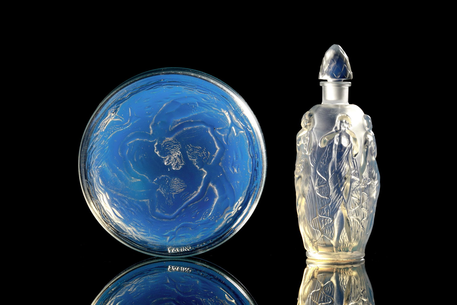 ART DECO FRENCH SABINO IRIDESCENT GLASS PIECES: Two opalescent art glass vanity pieces by Marius-Ernest Sabino, including "La Ronde Fleurie" scent bottle with moulded nude figures and a faceted stopper, signed to underside.Together with a covered d