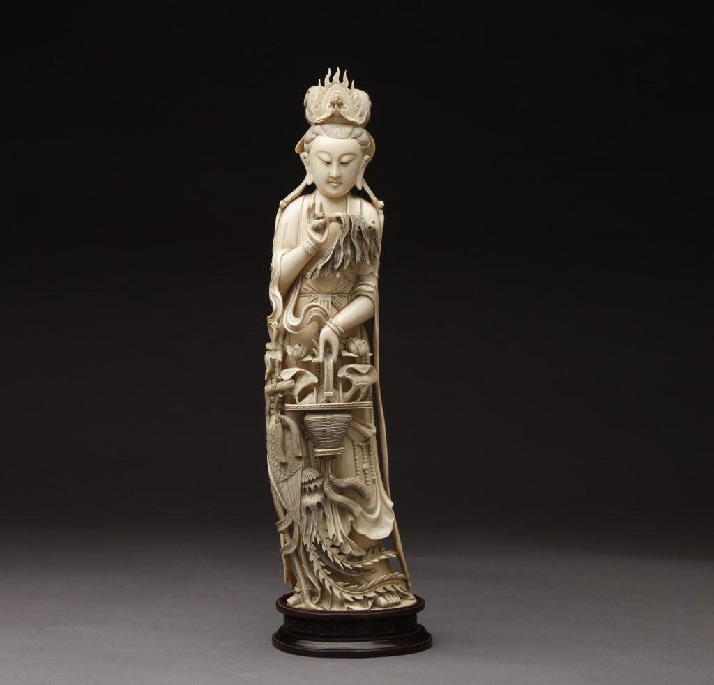 CHINESE LARGE CARVED IVORY FIGURE OF A GODDESS: The goddess is carved as holding a basket of fish in one hand with a crane besides her, a miniature Buddha is carved inside her halo. Late 19th century. H. 45.7cm (18 in.)