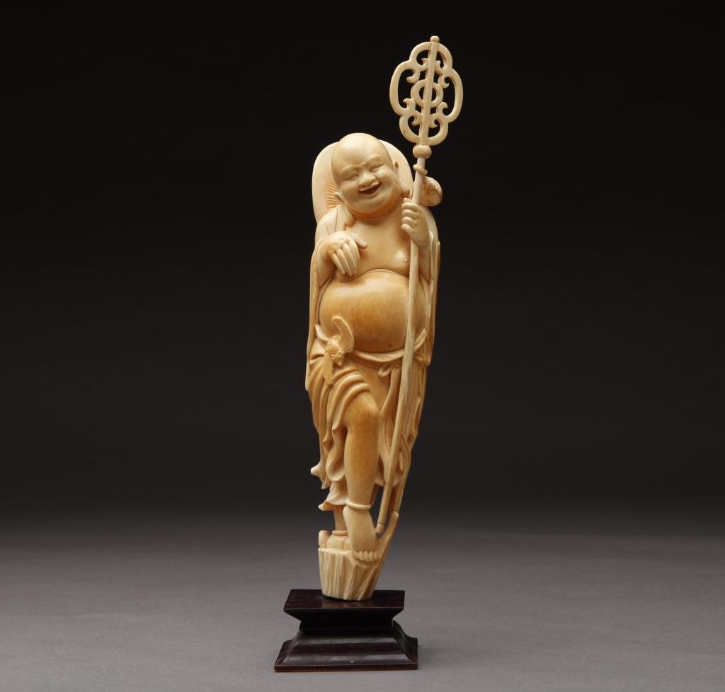 CHINESE IVORY CARVING OF BUDDHIST MONK: The monk has a shakujo in one hand and a bat next to him; attached to a wood stand. Late 19th century. H. without stand 33.3cm (13 1/8in.)