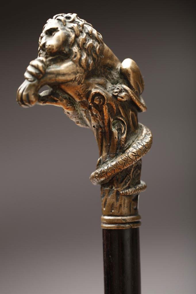 BRONZE LION-ON-SNAKE HANDLED WALKING STICK: Handle is formed as a lion crouching on a snake-wrapped tree trunk, on exotic wooden shaft, with horn ferrule. L. 89cm (35 in.)