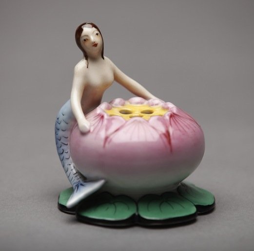 NORITAKE FIGURAL FLOWER FROG: Nude mermaid holding a pink and green lily pad with piercing for flowers. Red M-in-Wreath, H 8.5 cm. (3 3/8 in.)