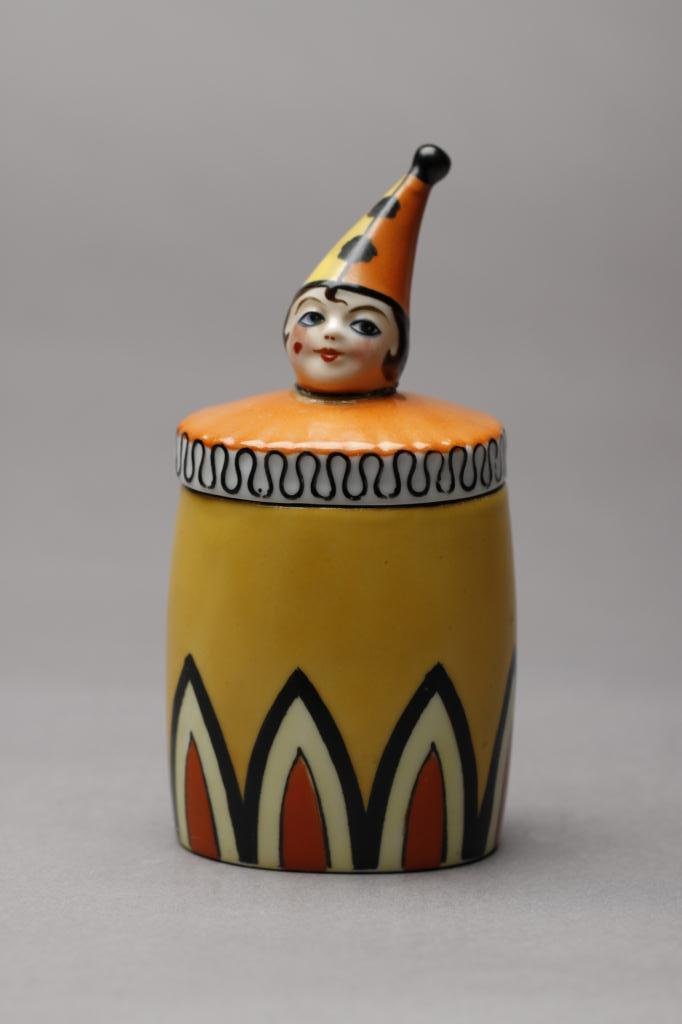 NORITAKE FIGURAL DRESSER JAR: Clown in an orange and yellow suit and cone cap (restoration to head), red M-in-Wreath, 29.1. H 14.5 cm. (5 3/4 in.)