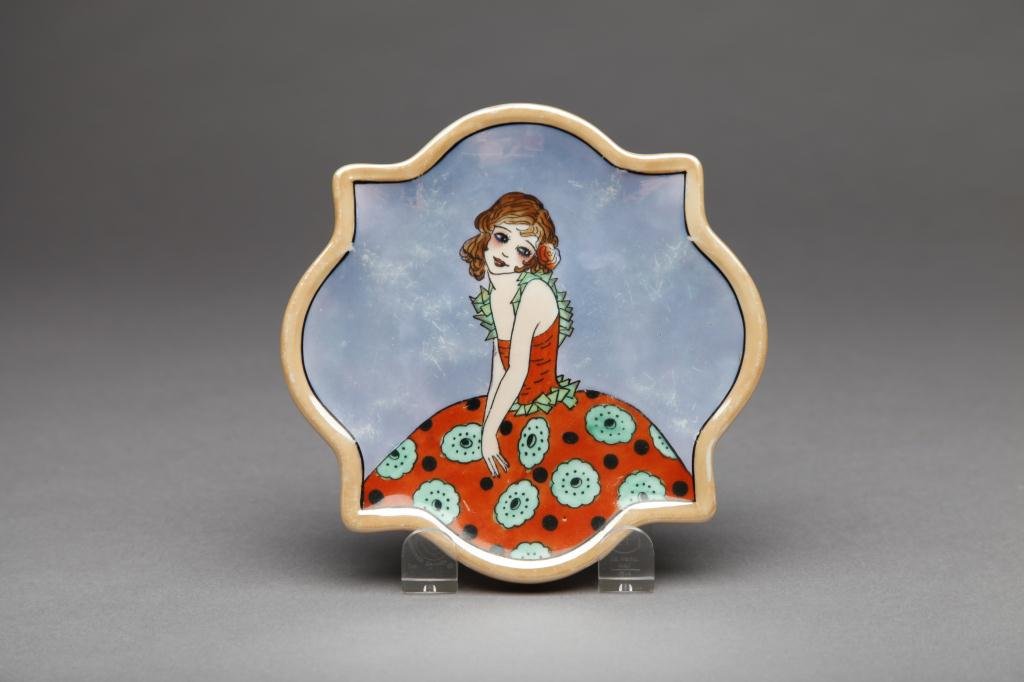 NORITAKE TILE / TEAPOT STAND: Scalloped body in orange and blue lustre with a young girl in a red and green dress (wear to blue). Green M-in-Wreath, 27.0. W 15.3 cm. (6 in.)