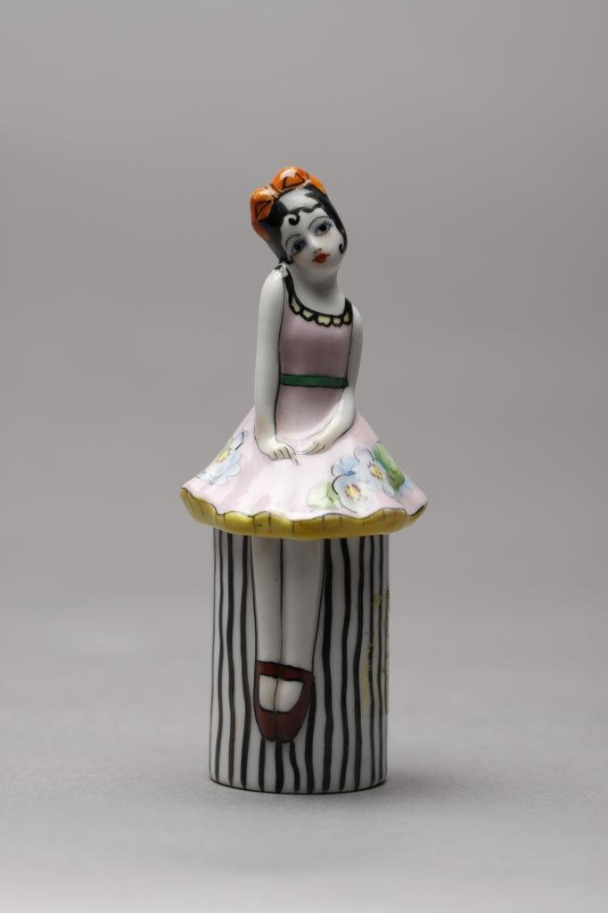 NORITAKE FIGURAL DRESSER JAR: Full body girl in pink dress sitting on a black and white stripe bench (minor chip to interior rim). Red M-in-Wreath, 27.3. H 12.2 cm. (4 3/4 in.)