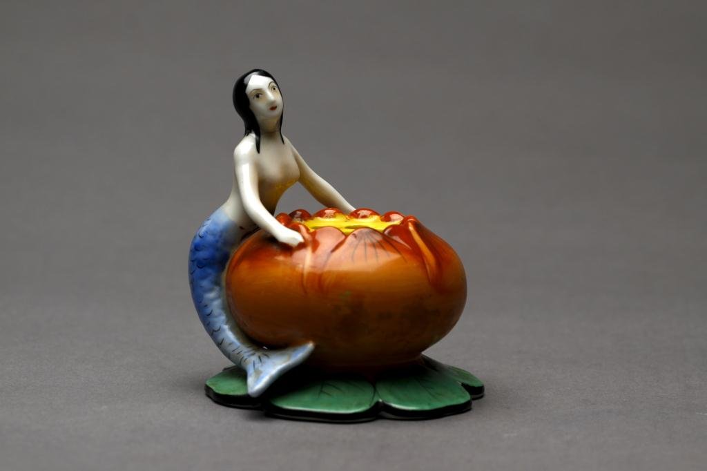 NORITAKE FIGURAL FLOWER FROG: Nude mermaid holding an orange and green lily pad with piercing for flowers. Green M-in-Wreath, H 8.8 cm. (3 1/2 in.)