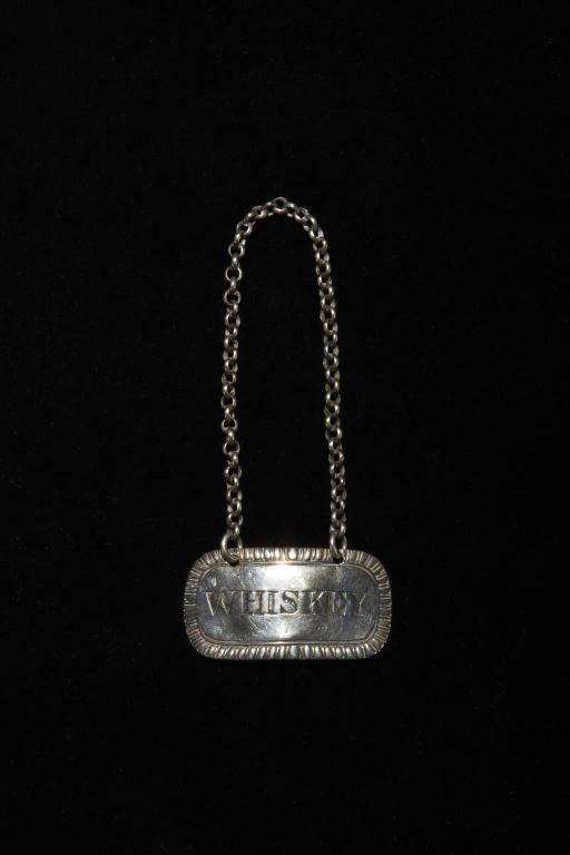 GEORGIAN SILVER BOTTLE TICKET: A George III sterling bottle ticket London 1812. Maker probably John Robins. Engraved on the front with WHISKEY and on the back the royal coat of arms within a garter. Length 4.7 cm