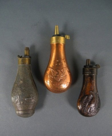 THREE GUN POWDER FLASKS: - copper eagle on a hummock pocket pistol flask H 10.16 cm. (4 in.), copper starr flask with brass sloping charger H 11.74 cm. (4 5/8 in.) and a zinc flask with embossed hunting scene marked PQ.F Pari