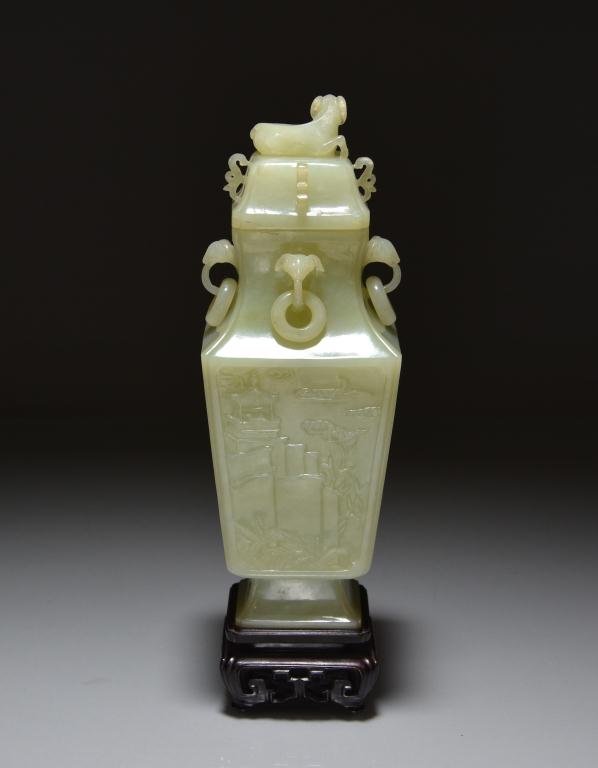 CHINESE CARVED CELADON JADE COVERED VASE: Of square form carved in low-relief with pagoda, pavilions, and riverside scene; neck has four lamp-head handles with mobile rings, with crouching lamp finial, on carved fitted wood stand. H. 22.5Cm (