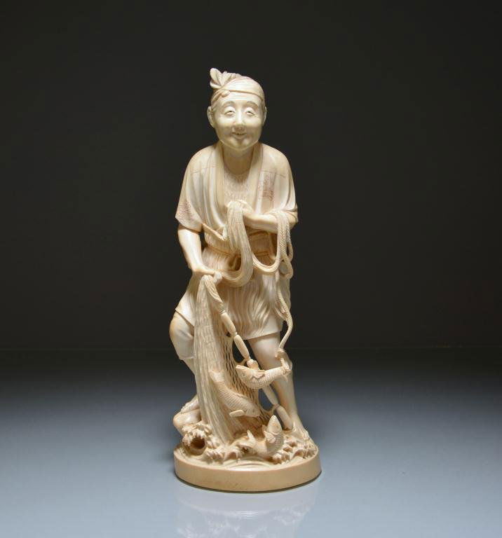 JAPANESE CARVED IVORY OKIMONO OF A FISHERMAN: Dressed in traditional clothes, harvesting fish with a net in one hand and holding a robe in the other; artist's mark to base, Japanese, Meiji. H. 32cm (12 5/8 in.) DISCLAIMER: This item is restricted
