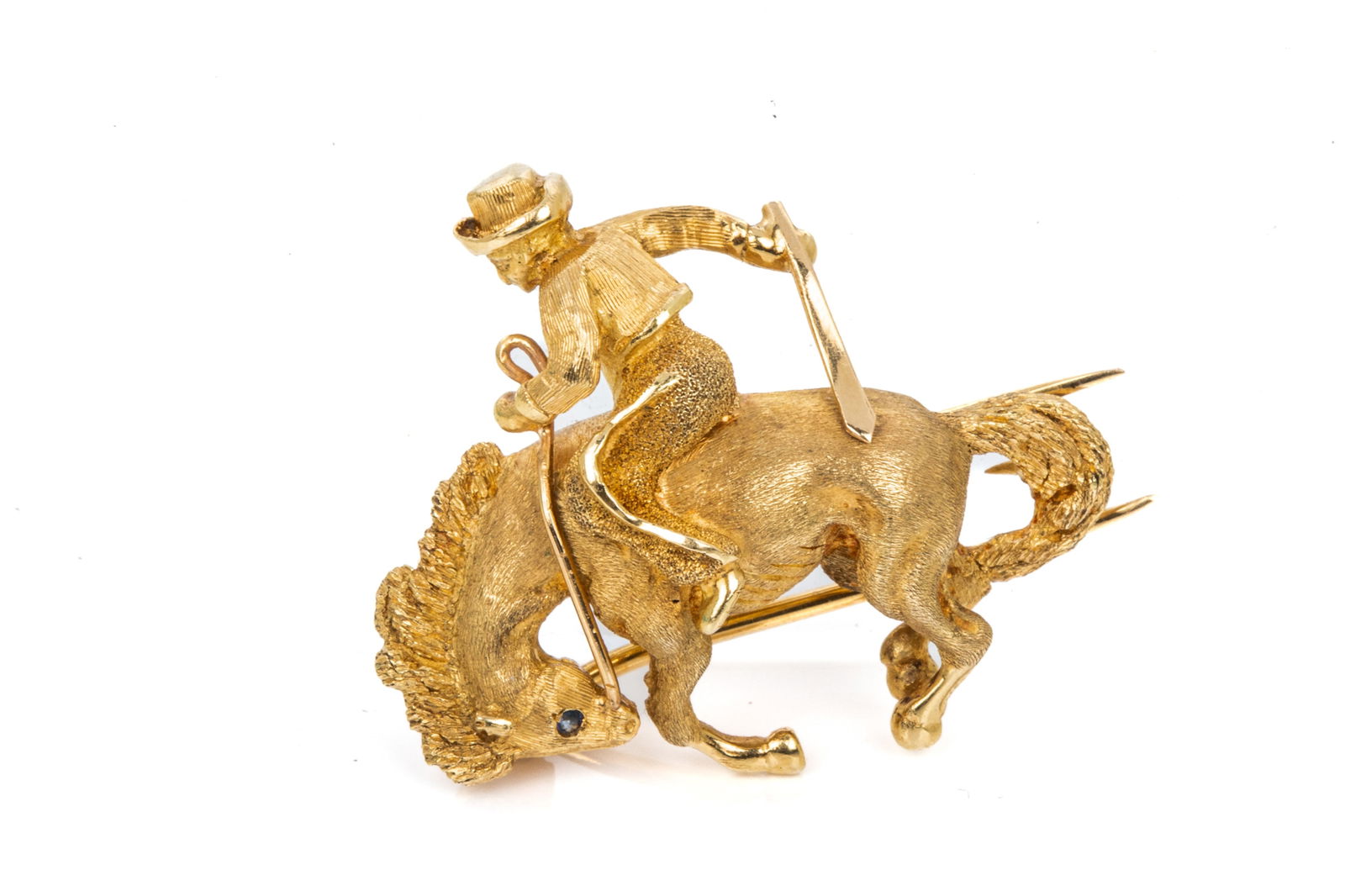 18K GOLD RODEO COWBOY PIN 8.1g: 18K yellow gold cowboy pin measuring 27.4mm x 30.2mm, equipped with spring-lock double pin closure with counter tooth. Featuring intricate detailing and a petite faceted sapphire eye. 8.1 grams