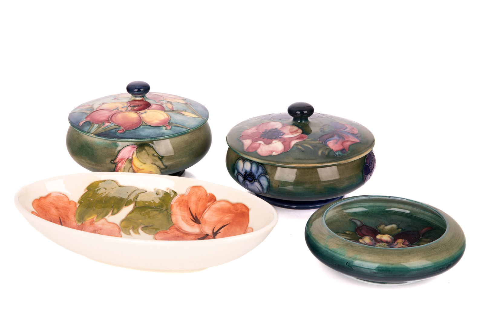 FOUR PCS OF MOORCROFT POTTERY: Two blue/green ground lidded bowls with floral motifs. One pale green to teal ombre ground ashtray in the "Columbine" pattern. One oval bowl with cream background in the "Hibiscus" pattern. 