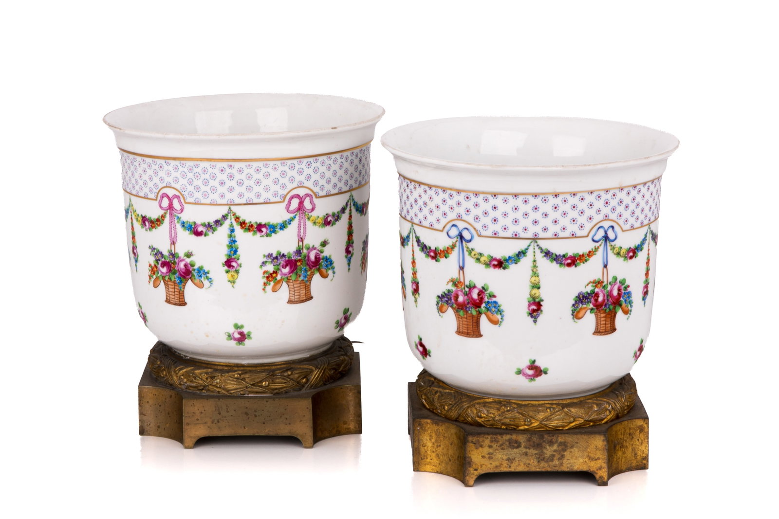 PAIR OF FRENCH PORCELAIN JARDINIERES: A pair of French Sevres-style porcelain jardinieres, featuring elegant white bodies decorated with hand-painted floral baskets and draping garlands. Each planter rests securely on ornate, decorative b