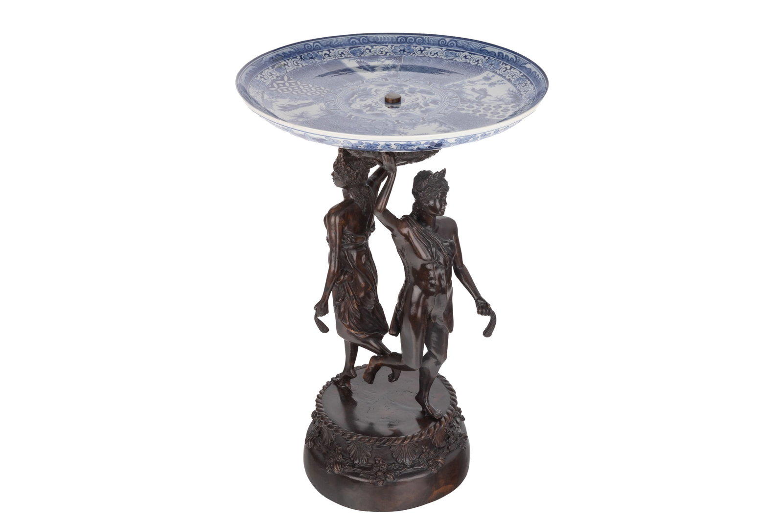 20TH C. BRONZE AND PORCELAIN TAZZA: A 20th century decorative bronze tazza centerpiece of two figures holding up a blue and white Japanese porcelain charger dish. H. 21 1/2in x 14 1/4in (54.5cm x 36.5cm)