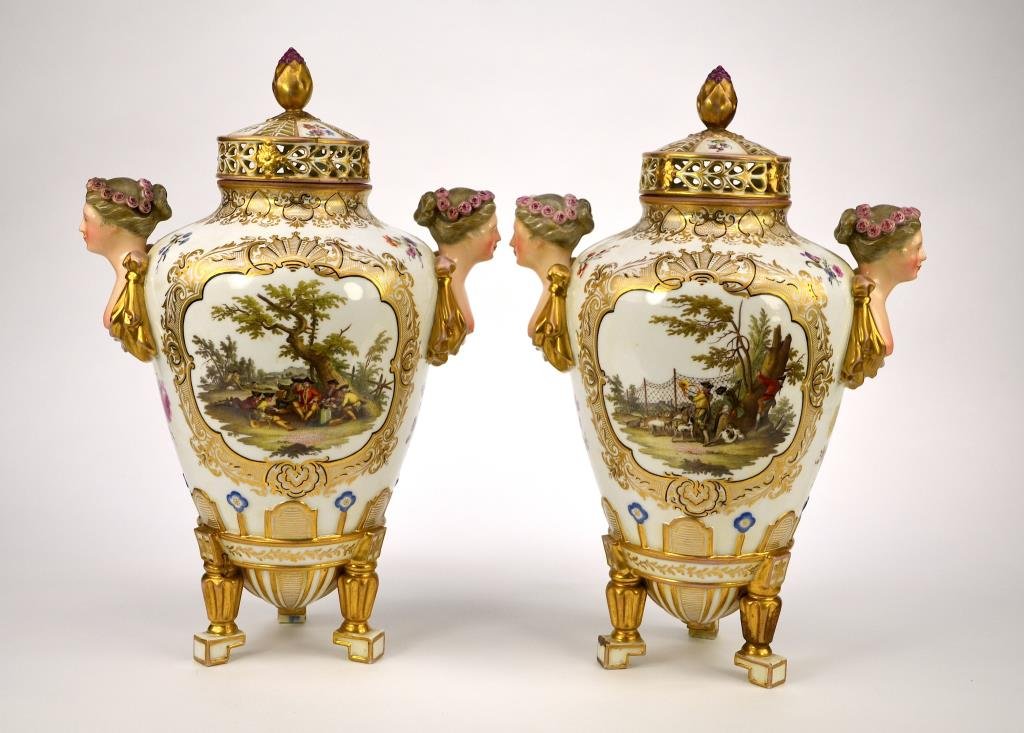 PAIR OF DRESDEN PORCELAIN ARMORIAL URNS: Painted on one side with two armorial crests and the other with a finely painted hunting scene surrounded by floral sprays, shoulders mounted with two nude busts raised on tripod feet, pierced covers.