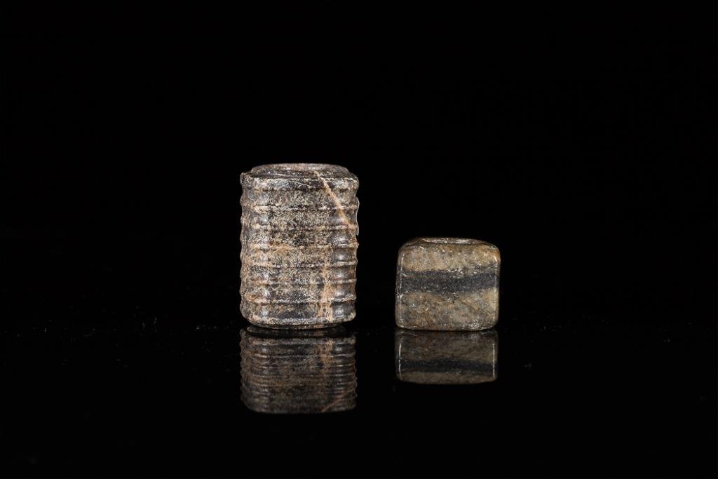 TWO NEOLITHIC STONE RITUAL CONG BEADS: The larger veined black and brown soapstone cong with a ridged body, the smaller mottled grey jade cong with square undecorated form. H of soapstone 5.3 cm. (2 1/8 in.) From the Irving Langleb collect