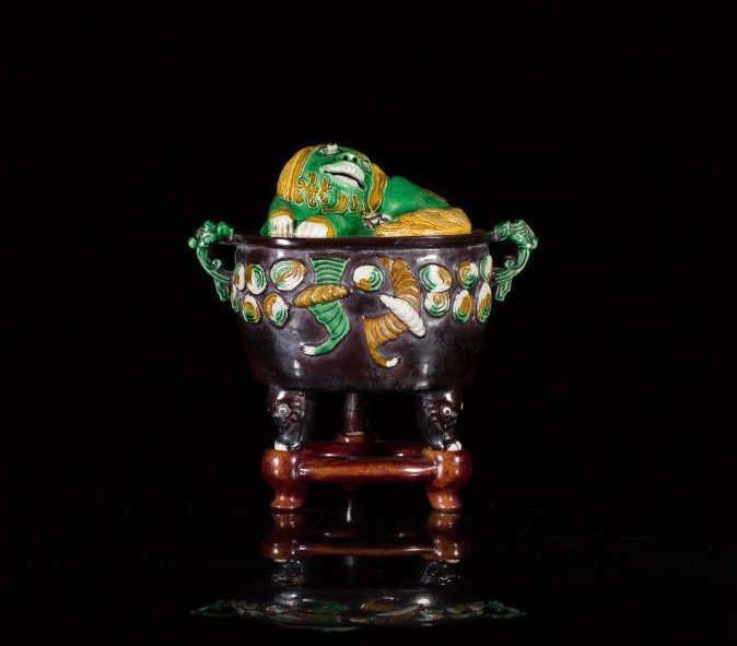 JAPANESE SANCAI GLAZED PORCELAIN TRIPOD CENSER: The dark-brown glazed body is decorated with bats and elephant-head handles, the cover as a fu-dog with mobile eyes. On carved fitted wood stand. 19th century or earlier. W. 19cm (7.5 in.)
