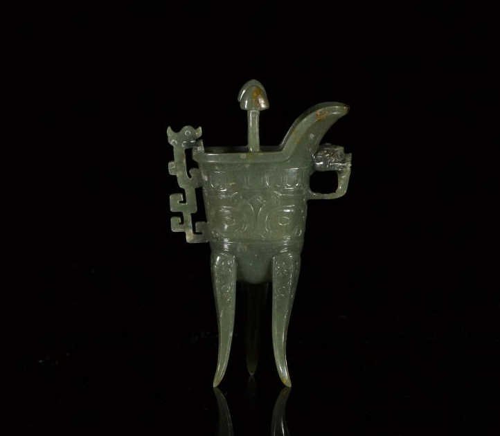 SMALL GREEN JADE ARCHAISTIC CHINESE WINE CUP, JUE: The body is carved with taotie masks under two spear-head posts, flanked by a qilin handle and a fu-dog loop under the spout, the three curved legs are further carved with ruyi-head pattern. H. 14.8cm