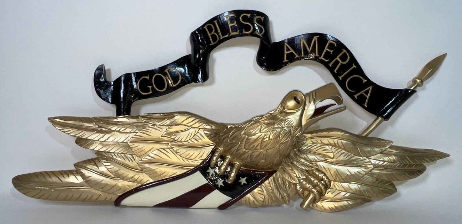 Carved Eagle with God Bless America Banner: Carved wood eagle with incised God Bless America banner and clutching a patriotic badge. Open beak with tongue. 4 lbs. 15" H x 33" W x 4" D.