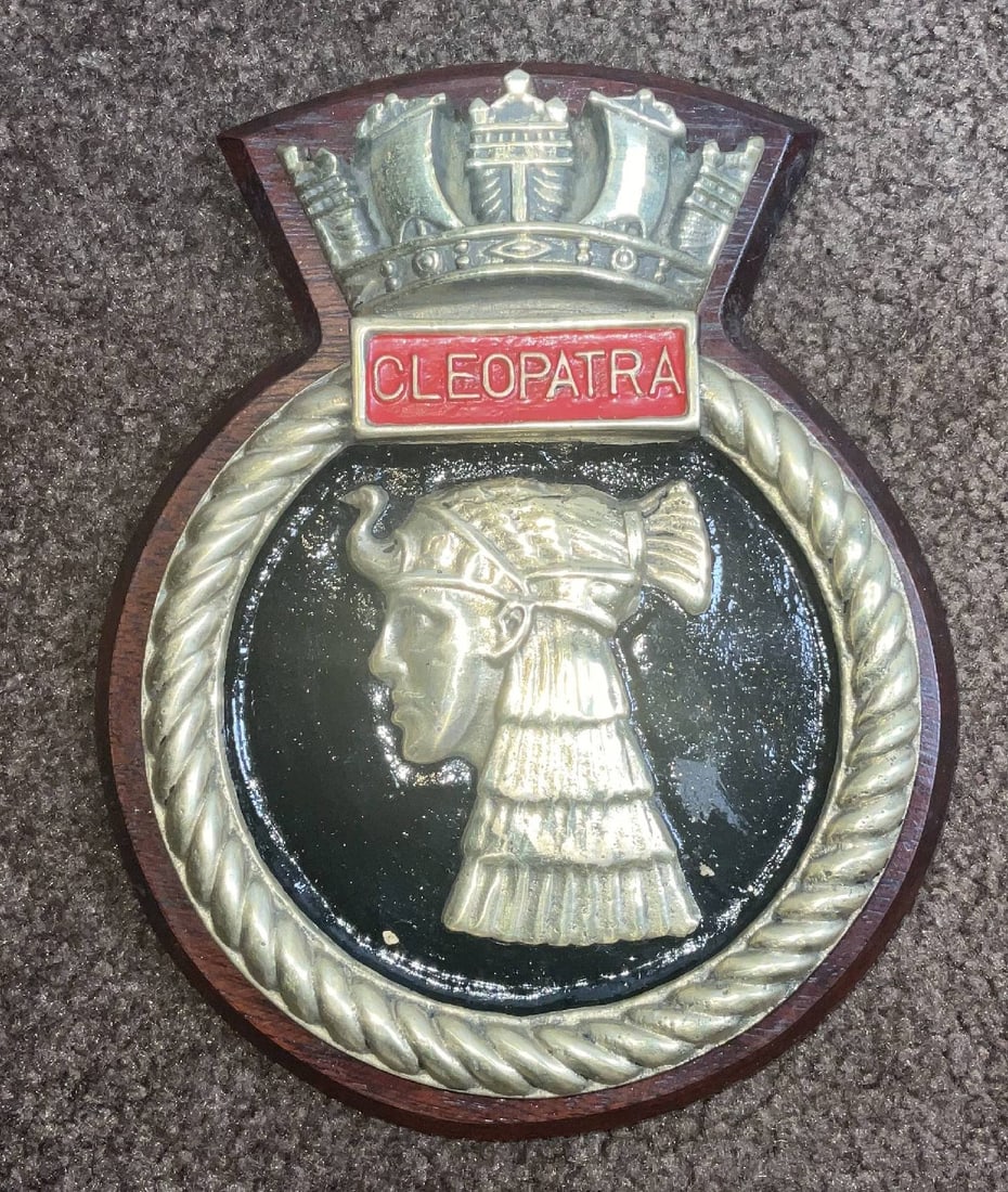 Royal Navy Ship's Badge from HMS Cleopatra: Ship's boat badge from HMS Cleopatra. Heavy brass tampion or badge with crown top. Mounted to a wood backboard. 6 lbs. 10" H x 8" W. HMS Cleopatra was a Dido-class cruiser of the Royal Navy. Launched 