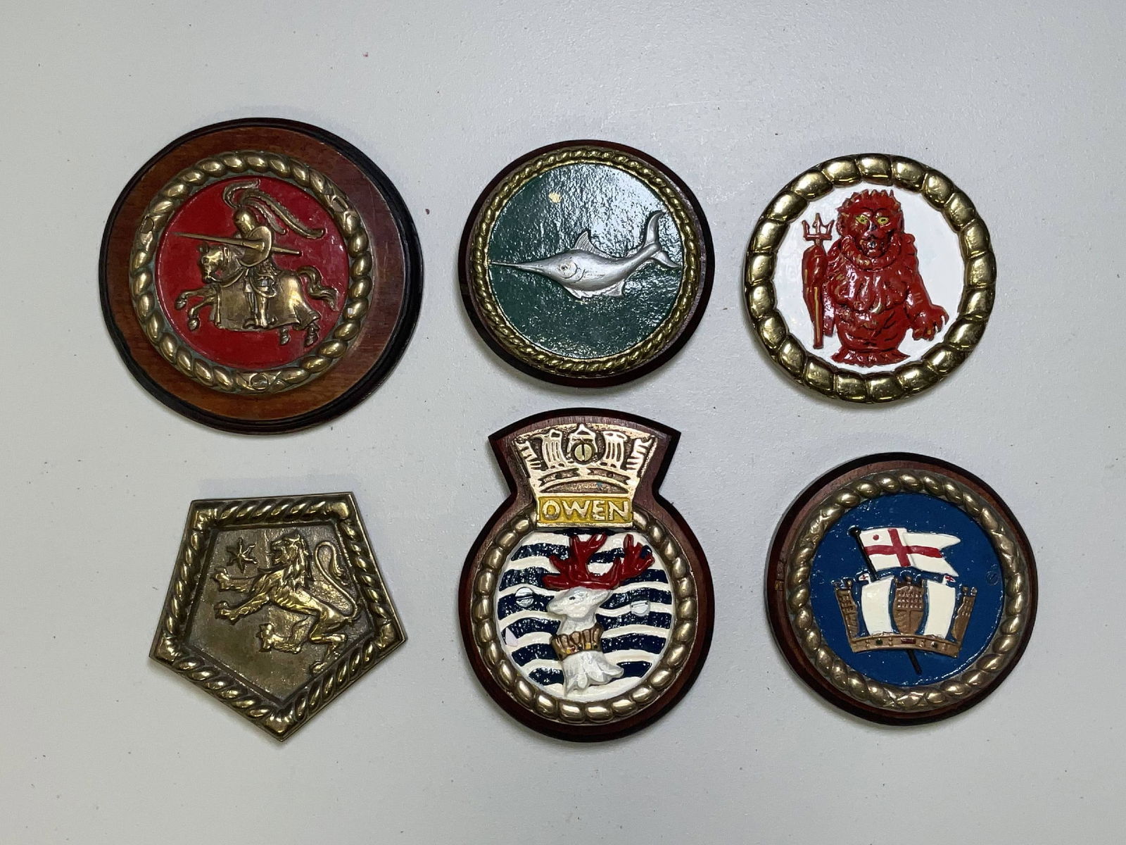 Royal Navy Gun Tompions and Badges from British Warships: Group of five gun tompions and a pentagonal crest, all cast in brass. Ships include HMS Flagstaff, the cruiser HMS Enterprise, HMS Bermuda, HMS Swordfish, HMS Resolution, and a Rear Admiral badge. Qua