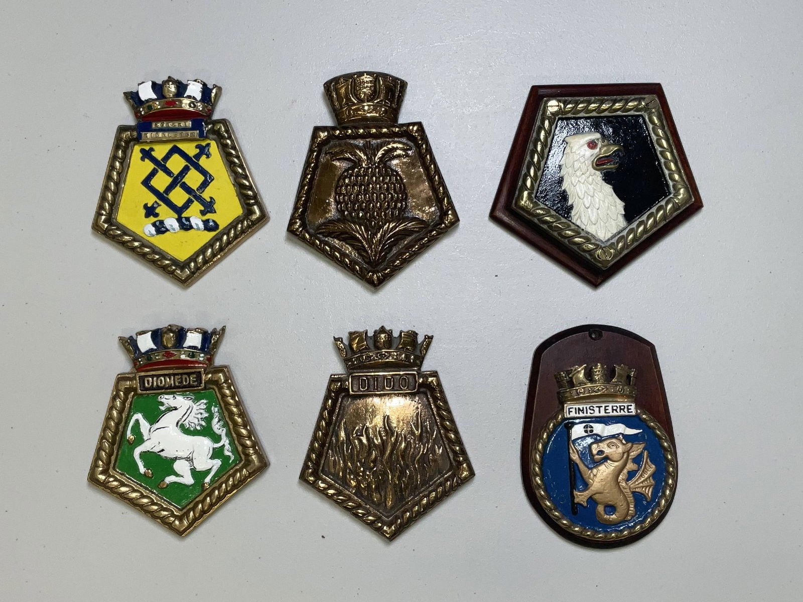 British Royal Navy Ships Badges from Various Warships: HMS Robert Middleton, a coastal stores carrier; HMS Jamaica, a Fiji class cruiser. HMS Diomede, a Danae class cruiser of 1938 built by Vickers, HMS Dido, a light cruiser and HMS Finistere. 17 lbs 6