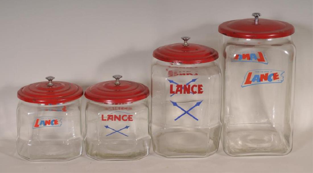 4 Lance Country Store Glass Display Jars: Four Lance glass display jars, each with red metal Lance lid; one approx. 8 1/2" x 7", 11"H; two approx. 7 1/2" x 6 1/2", 7 1/2"H; one approx. 8 1/2" x 7", 14"H; mid-size jar with chip to rim, others