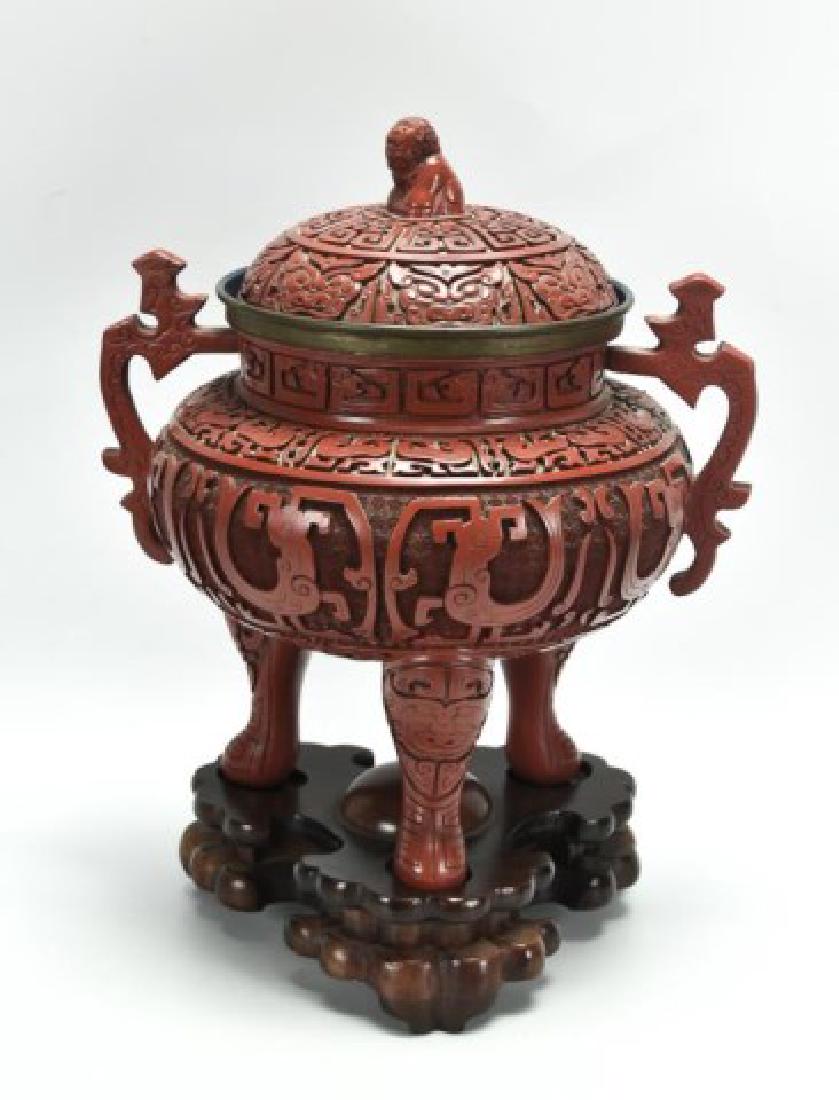 Large Chinese Faux Cinnabar Tripod Censer: Rounded body flanked by shaped handles, supported on three legs, domed lid with foo dog finial, overall decoration in molded relief, carved wood base; approx. 11 1/2"W, 14"H including 2 1/4"H stand