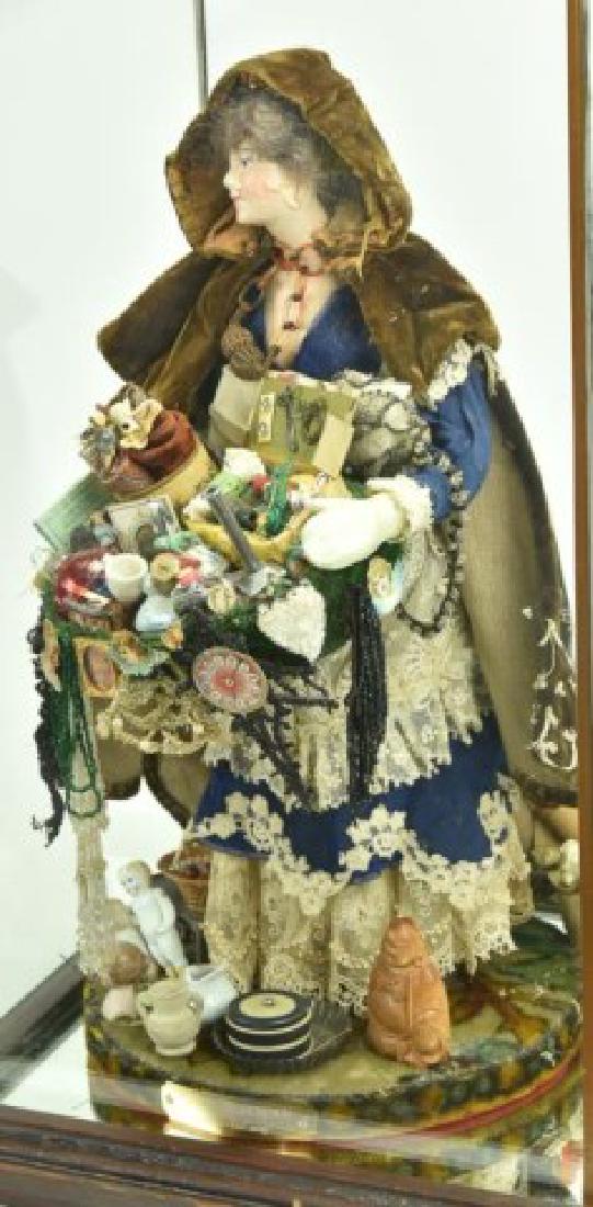 Norma Lizabeth DeCamp (North Carolina, 20th C.): Handcrafted original papier mache female street vendor doll under glass, wearing a lace trimmed dress under an embroidered and tattered brown velvet cape; holding a carton brimming with items for sale