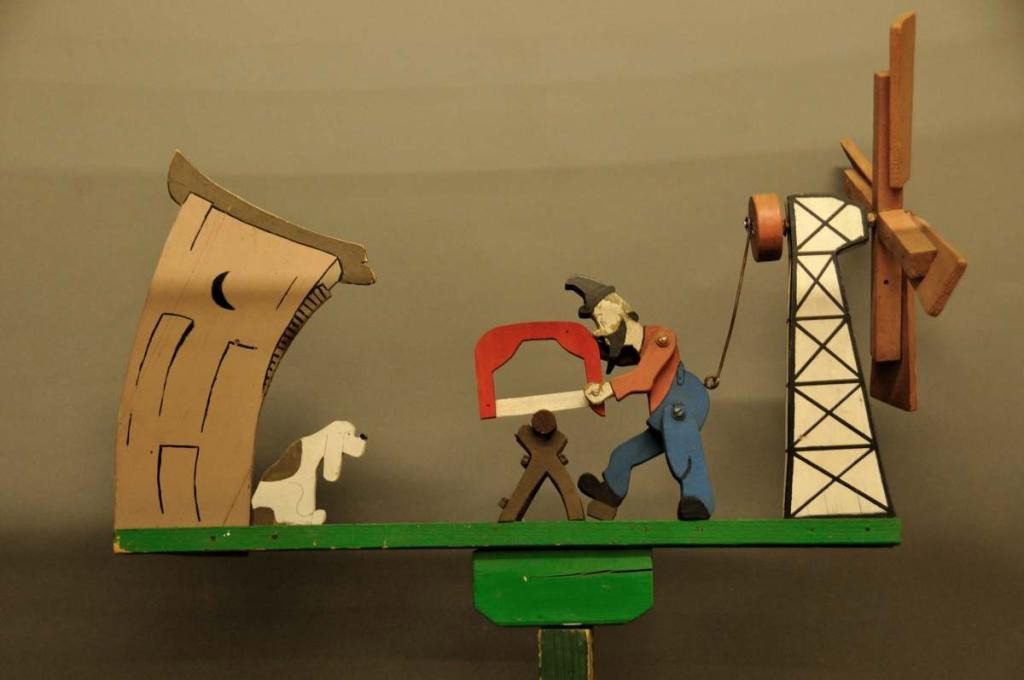 Painted Whirligig on Stand: Depicting a man sawing wood while his dog watches from in front of the outhouse; approximately 4'H, 23.5"W