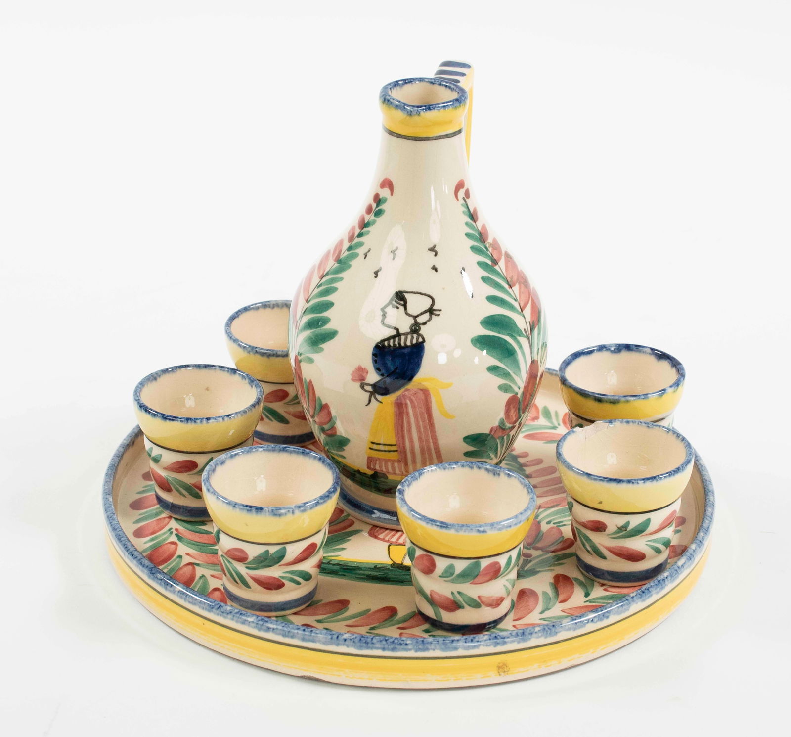 8-Piece Quimper Cordial Set: Pitcher, six cordial glasses, and round tray; 6"H pitcher and 8 1/4" round tray with typical peasant decoration; six 1 3/4"H glasses with floral and leafy bands; pitcher, tray and one glass marked on