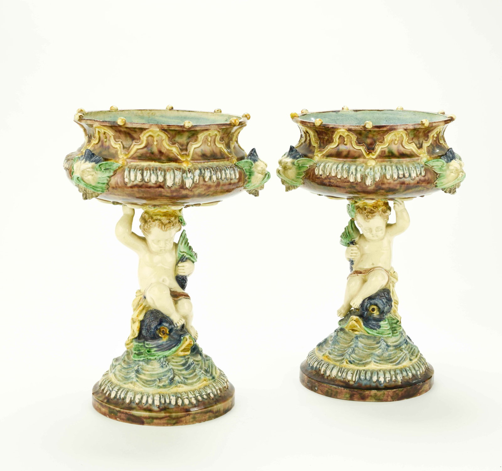 Pair of French Palissy Majolica Comports: Qualifies for in-house shipping---Each having a round bowl decorated with three masks, supported on a pedestal comprised of a putti riding a dolphin atop waves, round foot, each with four painted dots