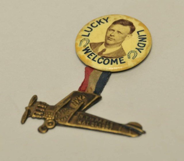 Charles Lindbergh Spirit of St. Louis Badge: Metal replica of Spirit of St. Louis embossed "N.Y. to Paris May 21, 1927" suspended by ribbon from circular badge with portrait of Lindbergh flanked each side by a horseshoe and reading "Lucky Lindy 