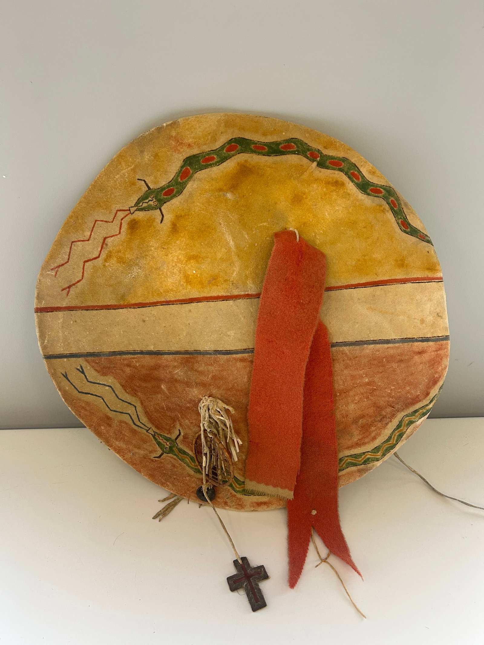 HANDCRAFTED PLAINS STYLE RAWHIDE SHIELD: APPROX: 19" DIAMETER, HAND PAINTED SERPENT AND LIGHTNING MOTIFS. RAWHIDE CORE WITH A SOFT HIDE WRAP. DREAMCATCHER, MEDICINE BUNDLE, FIBER CROSS, TRADE CLOTH, AND METAL CONE DECORATIONS. PROVENANCE: