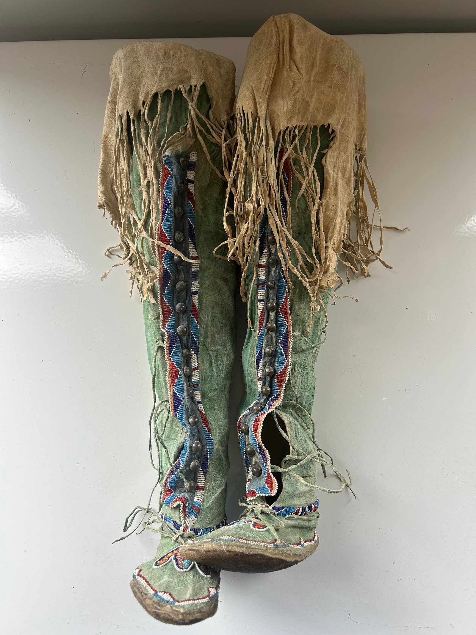 ANTIQUE SOUTHERN PLAINS BEADED WOMEN'S BOOTS: BEADED WITH GEOMETRIC DESIGN AND DECORATED WITH LEATHER FRINGE, BRASS BUTTONS AND GREEN PAINT.APPROXI. HEIGHT OF BOOT IS 25", LENGHT FROM TOE TO HEEL IS APPROX. 10" PROVENANCE: ESTATE OF DOUG DIVISH,