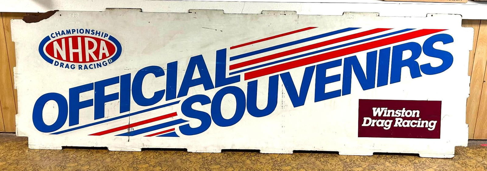 NHRA Drag Racing Official Souvenirs Masonite Sign: rated 7.75, 38"x119", the single-sided Masonite sign has good color, has wear and a few scratches, damage at the upper left-hand corner. We will not ship this lot.