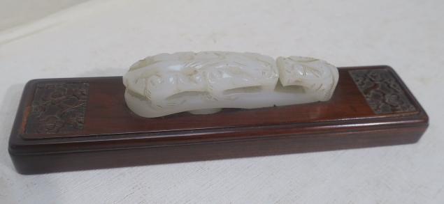 Carved Nephrite Jade Belt Hook on Rosewood Base:: Carved Nephrite Jade Belt Hook on Rosewood Base: Mutton Fat Jade Belt Hook is 3 3/4"L, Mounted on 7 1/4"L Rosewood Base with Floral Carved Ends. "The Old Dragon Teaching Its Young". (250-500)