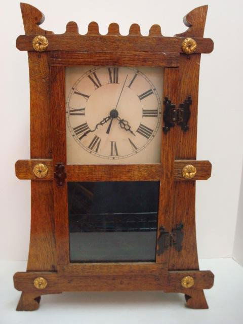 Oak Case Mission Style Shelf Clock: 230. Oak Case Mission Style Shelf Clock: Serpentine Hands with Roman Numerals on Paper Face. 13"W X 19 1/2"T. Hinged Glass Door with Metal Button Medallion Accents on Clock Front. Pendulum and Second