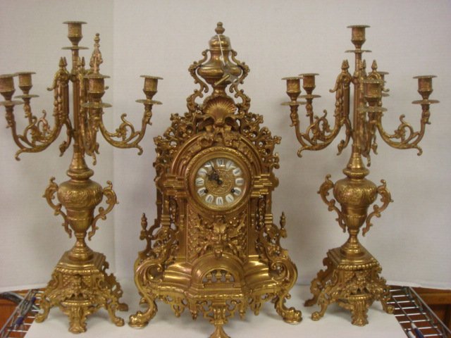 French Style Brass Garniture Set:: French Style Brass Garniture Set: Cast Brass Body with Elaborate Foliate Scroll Design and North Wind Mounted Cartouche. Matching 6 Light, 5 Arm Candelabra. German Movement Marked FHS (Franz Hermle) 1
