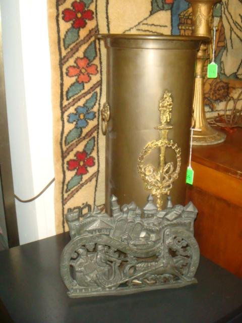 Brass Umbrella Stand and Sleigh Motif Trivet: Brass Umbrella Stand and Sleigh Motif Trivet: Umbrella Stand with Double Lion Head and Ring Handles and Applied Wreath and Torch on Front. 22 1/2"T. Molded 3 Legged Trivet with Cyrillic Writing is 13"