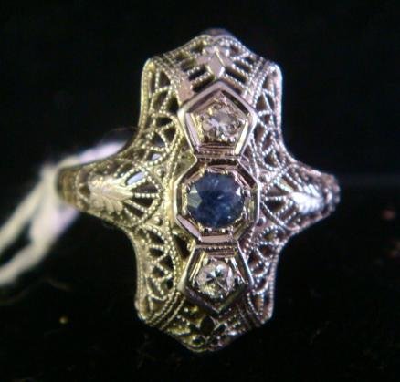 18K White Gold Ladies Deco Ring:: 18K White Gold Ladies Deco Ring: Classic Elongated Lacy Filigree Under Gallery Extending Down Shoulder. Prong Set Blue Sapphire Flanked Vertically by 2 Round Diamonds. Romantic Delicate Style. Size 5.