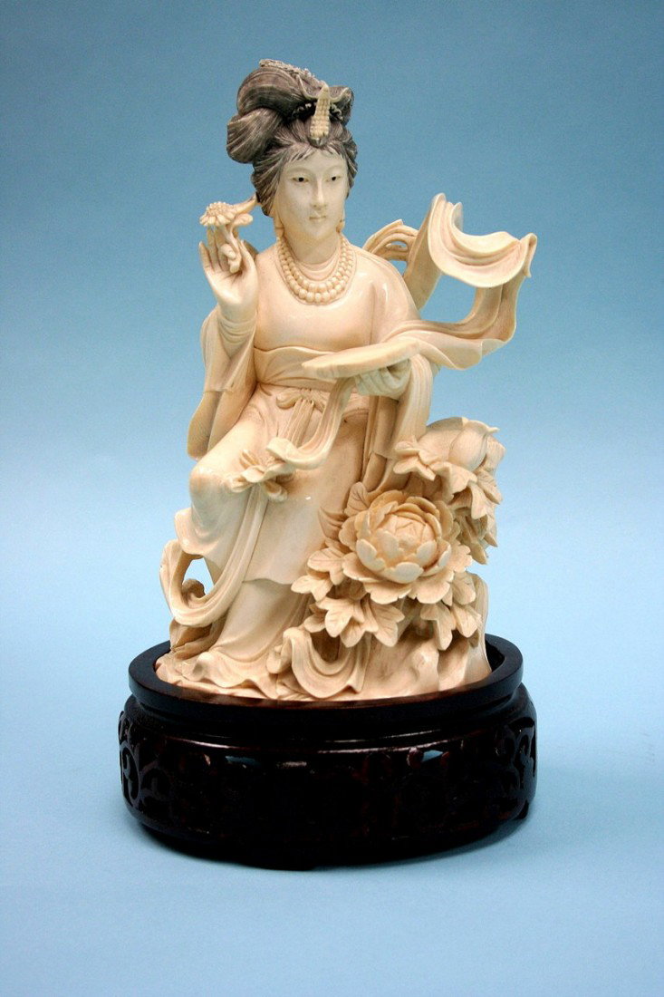 PEKING SCHOOL - A carved ivory figure of a Quan-Yi: PEKING SCHOOL - A carved ivory figure of a Quan-Yin seated on a rock and looking at a mirror in her hand. Well detailed carving. On pierce carved blackwood stand. 8 1/2" high.