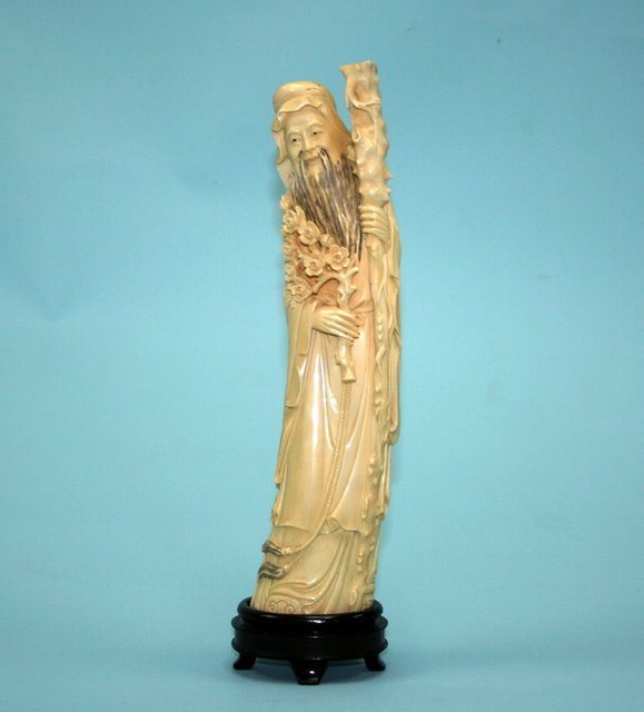 An early 20th century Chinese carved ivory figure: An early 20th century Chinese carved ivory figure of an old man supported by a large staff and holding peonies in his hand. Signed underside. On blackwood stand. 12" high.