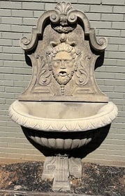 Cast stone wall fountain with a Bacchus head spitting water into a fluted basin with a tripod base.