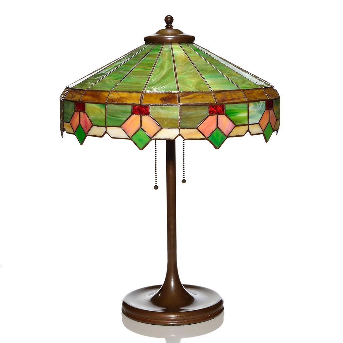 Arts & Crafts style leaded glass lamp, 16 1/4" shade: Arts & Crafts style leaded glass lamp. The 16 1/4 inch shade has a geometric drop design in pink and green, amber tortoise shell band and a green slag dome. The humble, bronze color metal base has two