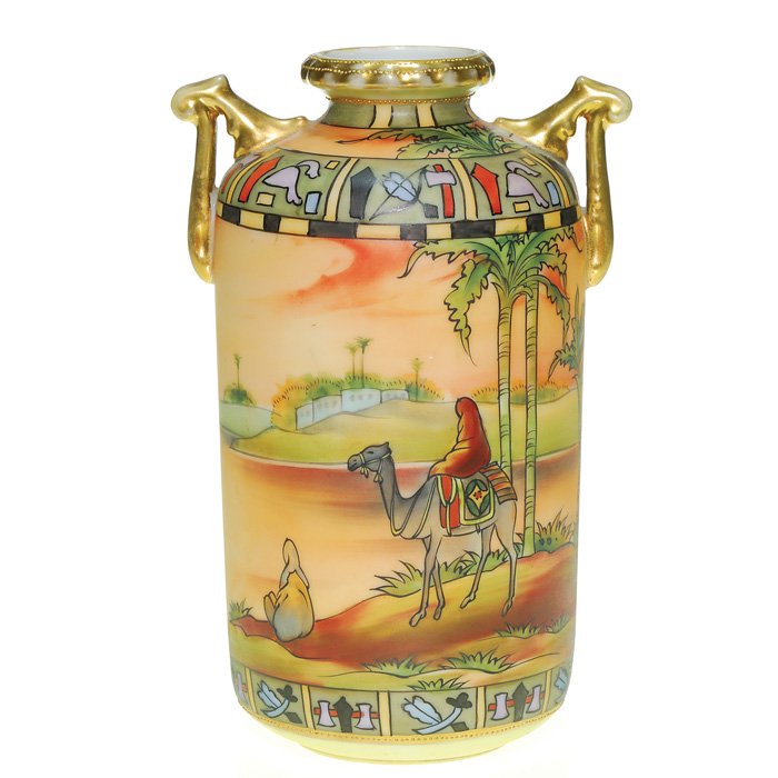 Nippon twig handled vase, Arabia scenic, 7 1/2”: Nippon mat finish vase showing a camel and rider strolling over a Middle Eastern landscape and beside a lake with palm trees and buildings on the horizon against a blazing orange sky. The shoulder and