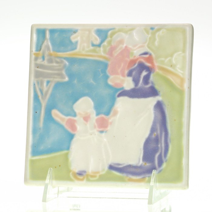 Rookwood Dutch 5 3/4” trivet, #3203, 1926: Trivet depicting a Dutch woman holding her smaller child while a daughter stands by her side, waving to a boat in the harbor with a windmill in the background, made in 1926 and colored in glazes of iv