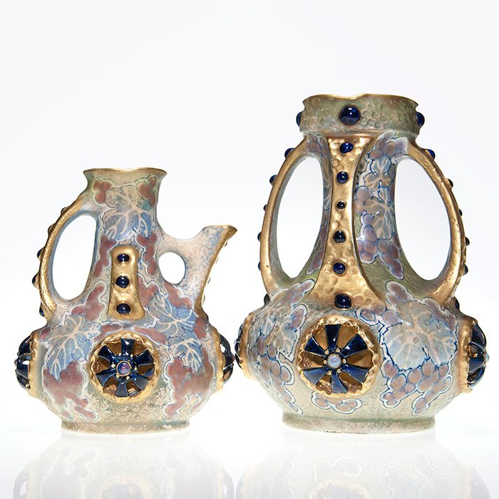 2 Amphora pieces, Pitcher, Vase, 9 1/2 + 7 1/4”: Two Amphora pieces with the same style of decoration. 1. Pitcher with overall grape decor along with 4 reticulated flower heads trimmed with fired on gold standing 7 1/4 inches tall. 2. Vase with same